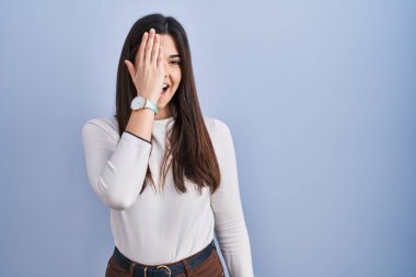 Mavi arka planda duran genç esmer kadın bir gözünü eliyle kapatıyor, yüzünde kendine güvenen bir gülümseme ve sürpriz bir duygu var.. 