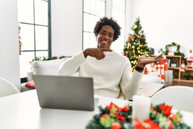 Genç Afro-Amerikalı adam dizüstü bilgisayar kullanıyor Noel ağacının yanında hayrete düşmüş ve kameraya gülümsüyor.. 
