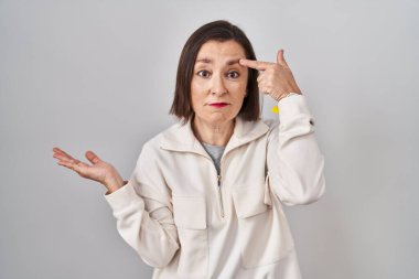 Middle age hispanic woman standing over isolated background confused and annoyed with open palm showing copy space and pointing finger to forehead. think about it. 