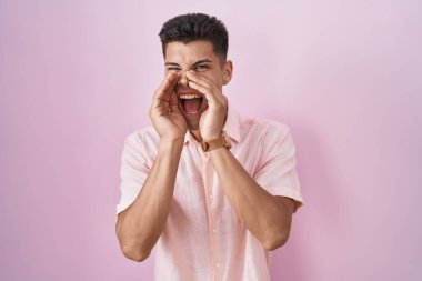 Young hispanic man standing over pink background shouting angry out loud with hands over mouth 