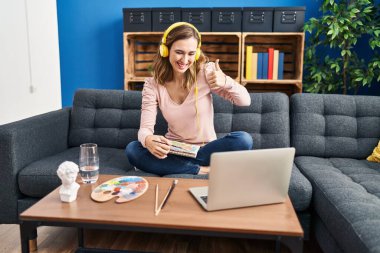 Young beautiful woman practicing painting looking at online tutorial smiling happy and positive, thumb up doing excellent and approval sign 