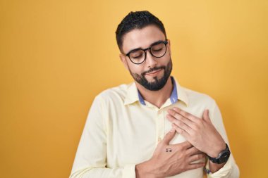 İspanyol genç adam iş kıyafetleri ve gözlük takıyor elleri göğsünde, gözleri kapalı gülümsüyor ve yüzünde minnet ifadesi var. sağlık kavramı. 