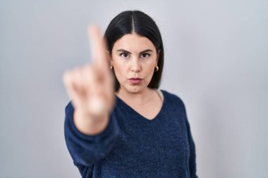 Genç esmer kadın izole edilmiş arka planda duruyor. Parmağını kaldırmış ve kızgın bir ifadeyle işaret ediyor. 