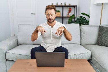 Middle age man using laptop at home pointing down looking sad and upset, indicating direction with fingers, unhappy and depressed. 
