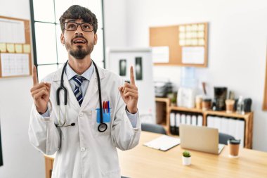 Hispanic man with beard wearing doctor uniform and stethoscope at the office amazed and surprised looking up and pointing with fingers and raised arms. 