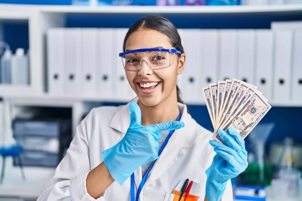 Young brazilian woman working at scientist laboratory holding money smiling happy pointing with hand and finger