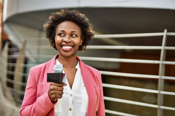 Young african american woman journalist holding reporter microphone speaking and smiling to the camera for television news