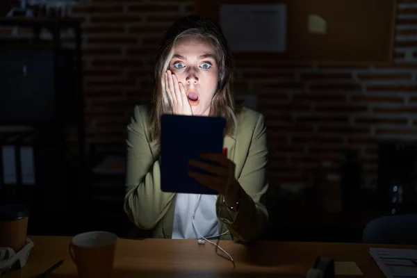 Blonde caucasian woman working at the office at night afraid and shocked, surprise and amazed expression with hands on face