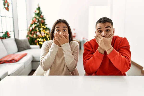 他那对惊慌失措的年轻夫妇坐在圣诞树旁的桌子上 双手捂着嘴 为自己的错误而震惊 秘密概念 — 图库照片