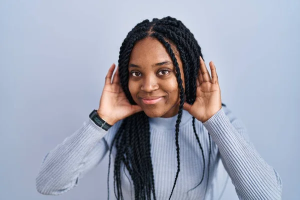 African american woman standing over blue background trying to hear both hands on ear gesture, curious for gossip. hearing problem, deaf