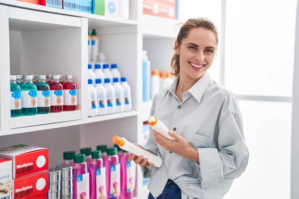 Young woman customer smiling confident holding sunscreen bottles at pharmacy