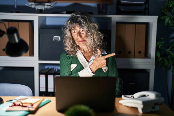Middle age woman working at night using computer laptop pointing aside worried and nervous with forefinger, concerned and surprised expression