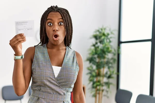 Young african american woman with braids holding covid record card scared and amazed with open mouth for surprise, disbelief face