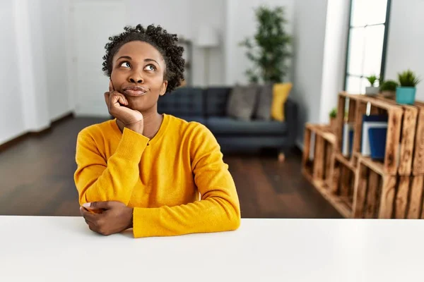 Young african american woman wearing casual clothes sitting on the table at home with hand on chin thinking about question, pensive expression. smiling with thoughtful face. doubt concept.