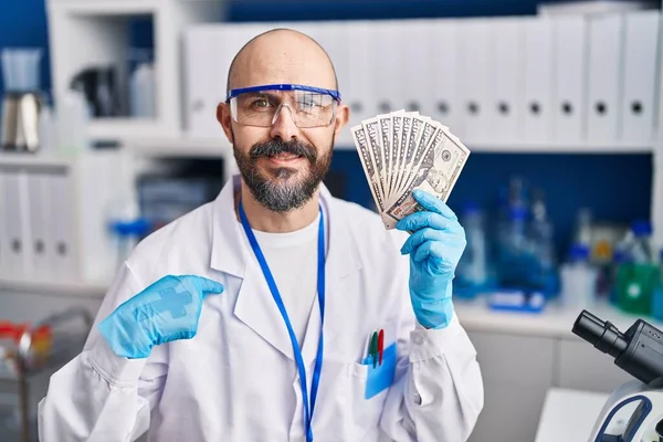 Young hispanic man working at scientist laboratory holding money pointing finger to one self smiling happy and proud