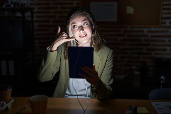 Blonde caucasian woman working at the office at night smiling doing phone gesture with hand and fingers like talking on the telephone. communicating concepts.