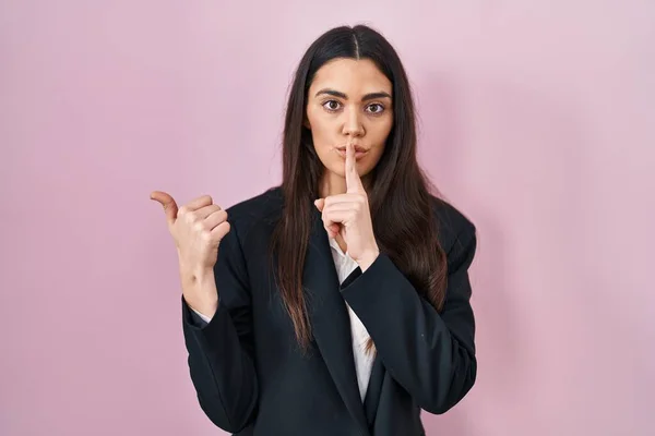 ピンクの背景の上にビジネススタイルを身に着けている若いブルネットの女性は 側に手で指して唇に指で静かになるように求めている 沈黙と秘密の概念 — ストック写真