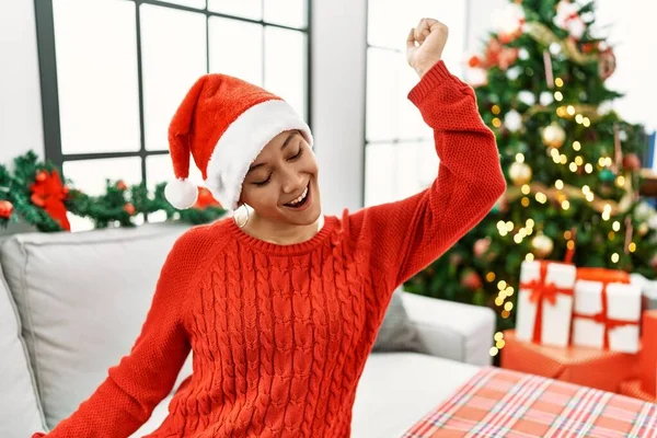 Young hispanic woman with short hair wearing christmas hat sitting on the sofa dancing happy and cheerful, smiling moving casual and confident listening to music