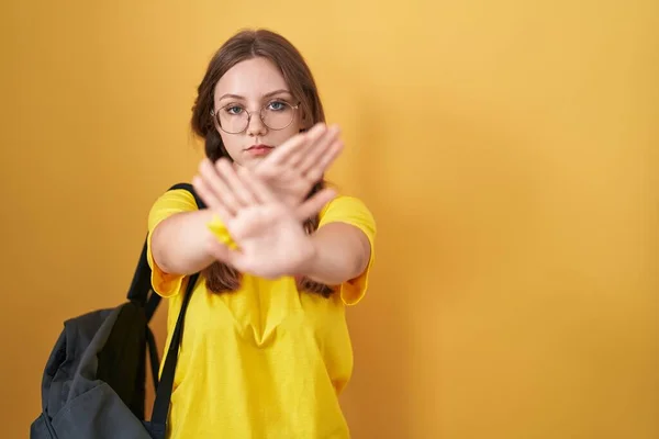 Young caucasian woman wearing student backpack over yellow background rejection expression crossing arms and palms doing negative sign, angry face