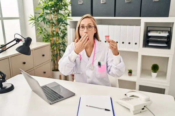 Young blonde doctor woman holding menstrual cup covering mouth with hand, shocked and afraid for mistake. surprised expression