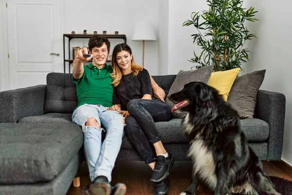Young caucasian couple watching tv sitting on the sofa with dog at home.