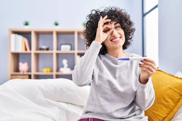 Young brunette woman with curly hair holding pregnancy test result smiling happy doing ok sign with hand on eye looking through fingers 