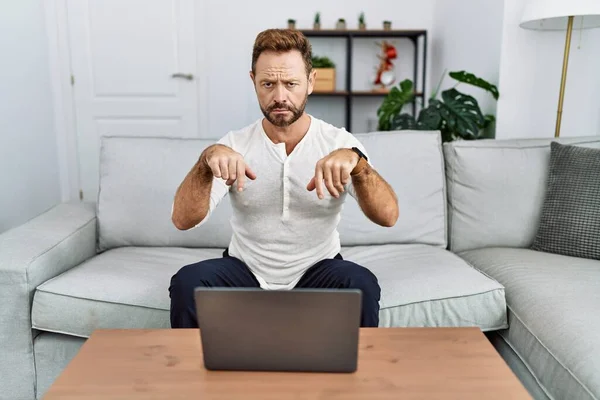 Middle age man using laptop at home pointing down looking sad and upset, indicating direction with fingers, unhappy and depressed.