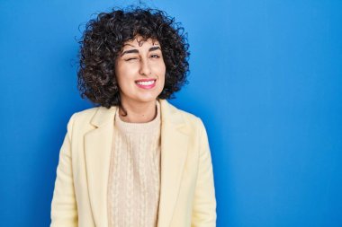 Young brunette woman with curly hair standing over blue background winking looking at the camera with sexy expression, cheerful and happy face. 