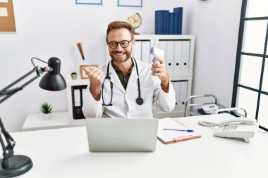 Middle age doctor man holding thermometer at the clinic pointing thumb up to the side smiling happy with open mouth 