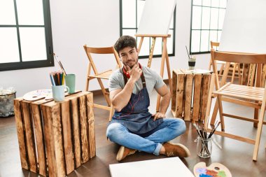 Young hispanic man sitting at art studio looking confident at the camera with smile with crossed arms and hand raised on chin. thinking positive. 
