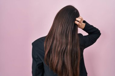 Genç esmer kadın pembe arka planda iş stili giyinmiş. Elleri havada şüpheyi düşünüyor. 