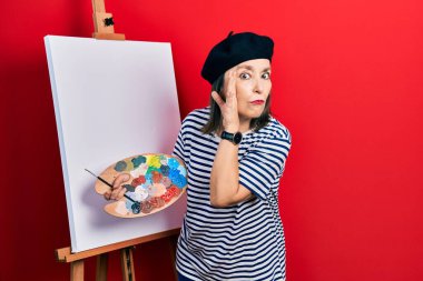 Middle age hispanic woman standing drawing with palette by painter easel stand hand on mouth telling secret rumor, whispering malicious talk conversation 