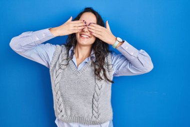 Genç esmer kadın mavi arka planda duruyor. Gözleri kaplıyor, neşeli ve komik gülümsüyor. kör kavram. 