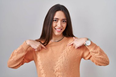 Genç esmer kadın beyaz arka planda duruyor. Kendine güveni tam görünüyor. Kendini gururlu ve mutlu bir şekilde gösteriyor.. 
