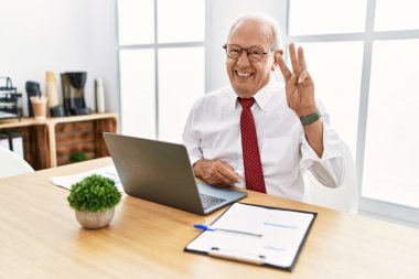 Senior man working at the office using computer laptop showing and pointing up with fingers number three while smiling confident and happy. 