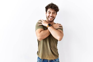 Hispanic man standing over isolated white background hugging oneself happy and positive, smiling confident. self love and self care 