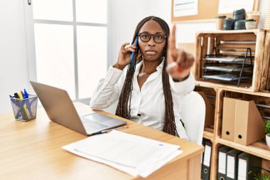 Black woman with braids working at the office speaking on the phone pointing with finger up and angry expression, showing no gesture 