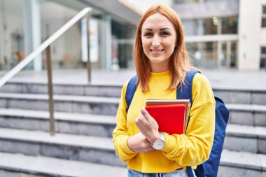 Üniversitede elinde kitaplarla gülümseyen beyaz bir bayan öğrenci.
