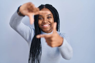 African american woman standing over blue background smiling making frame with hands and fingers with happy face. creativity and photography concept. 