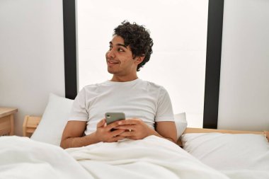 Genç İspanyol adam yatak odasındaki yatakta akıllı telefon kullanıyor..