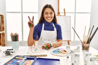 Resim stüdyosundaki genç esmer kadın boyalı elleriyle iki numarayı gösterip işaret ederken kendinden emin ve mutlu bir şekilde gülümsüyor.. 