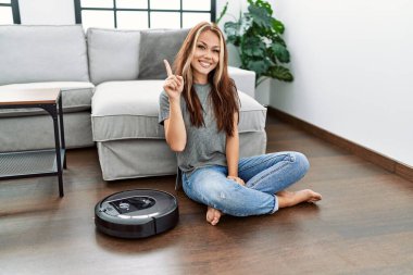 Young caucasian woman sitting at home by vacuum robot smiling happy pointing with hand and finger to the side 
