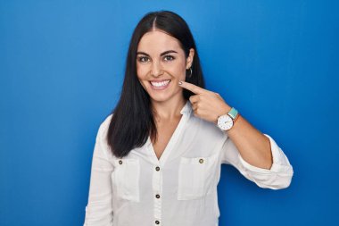 Mavi arka planda dikilen genç İspanyol kadın, parmakları burnuna ve yüzüne dönük, neşeli bir şekilde gülümsüyor. güzellik konsepti 