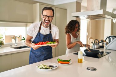 Middle age couple cooking mediterranean food at home smiling and laughing hard out loud because funny crazy joke. 