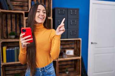 Akıllı telefon kullanan genç esmer kadın gülümsüyor mutlu bir şekilde elini ve parmağını yana doğru gösteriyor. 