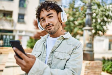 Yakışıklı, sakallı, güneşli bir günde mutlu bir şekilde gülümseyen kulaklık takan akıllı telefonlu müzik dinleyen bir İspanyol.