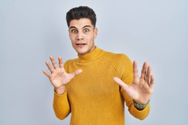 Young hispanic man standing over blue background afraid and terrified with fear expression stop gesture with hands, shouting in shock. panic concept. 
