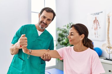 Orta yaşlı erkek ve kadın gülümsüyor kollarını esneterek fizik tedavi kliniğinde rehabilitasyon seansı yapıyorlar.