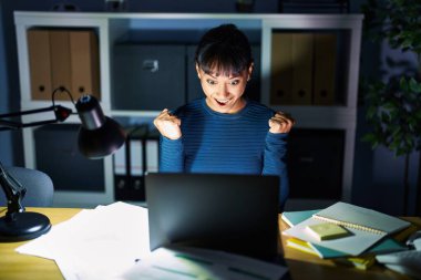 Geceleri ofiste çalışan genç ve güzel bir kadın kollarını kaldırıp, gözlerini açarak başarının şaşkınlığını ve şaşkınlığını yaşıyor. Kazanan konsept. 