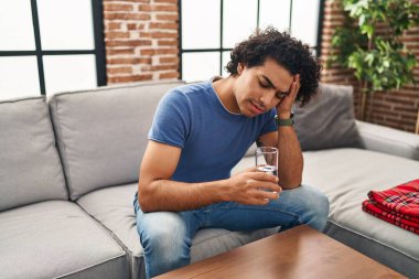 Genç İspanyol adam evdeki kanepede oturmuş su içiyor.